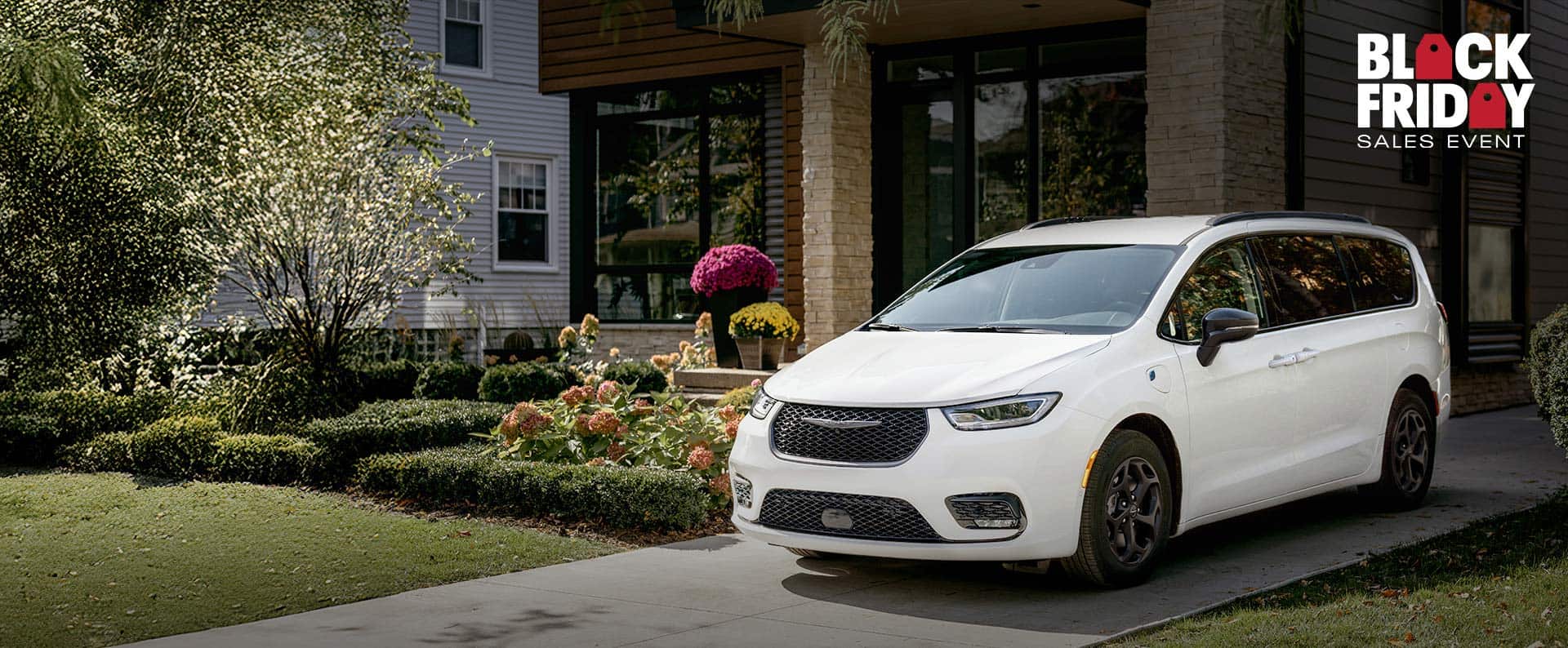 A driver-side front angle of a white 2025 Chrysler Pacifica Select Plug-in Hybrid with the Premium S Appearance Package, parked in the driveway of a neighborhood home. Black Friday Sales Event.
