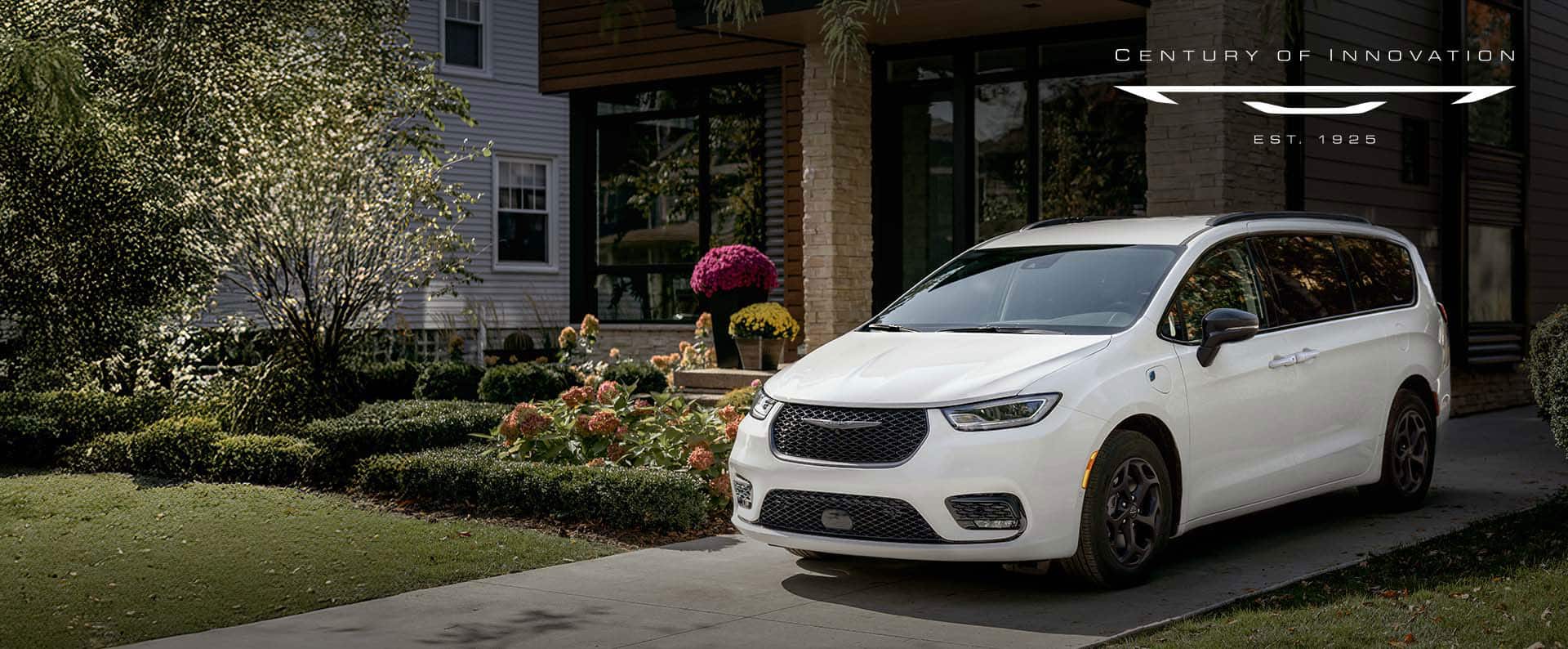 A driver-side front angle of a white 2025 Chrysler Pacifica Select Plug-in Hybrid with the Premium S Appearance Package, parked in the driveway of a neighborhood home. Century of Innovation.