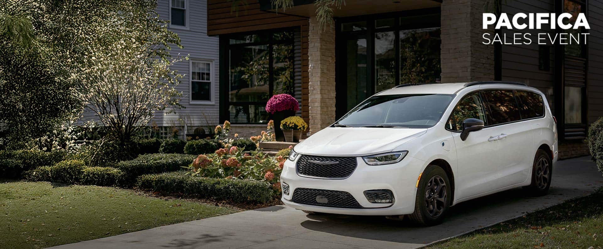 A driver-side front angle of a white 2025 Chrysler Pacifica Select Plug-in Hybrid with the Premium S Appearance Package, parked in the driveway of a neighborhood home. Chrysler Pacifica sales event.