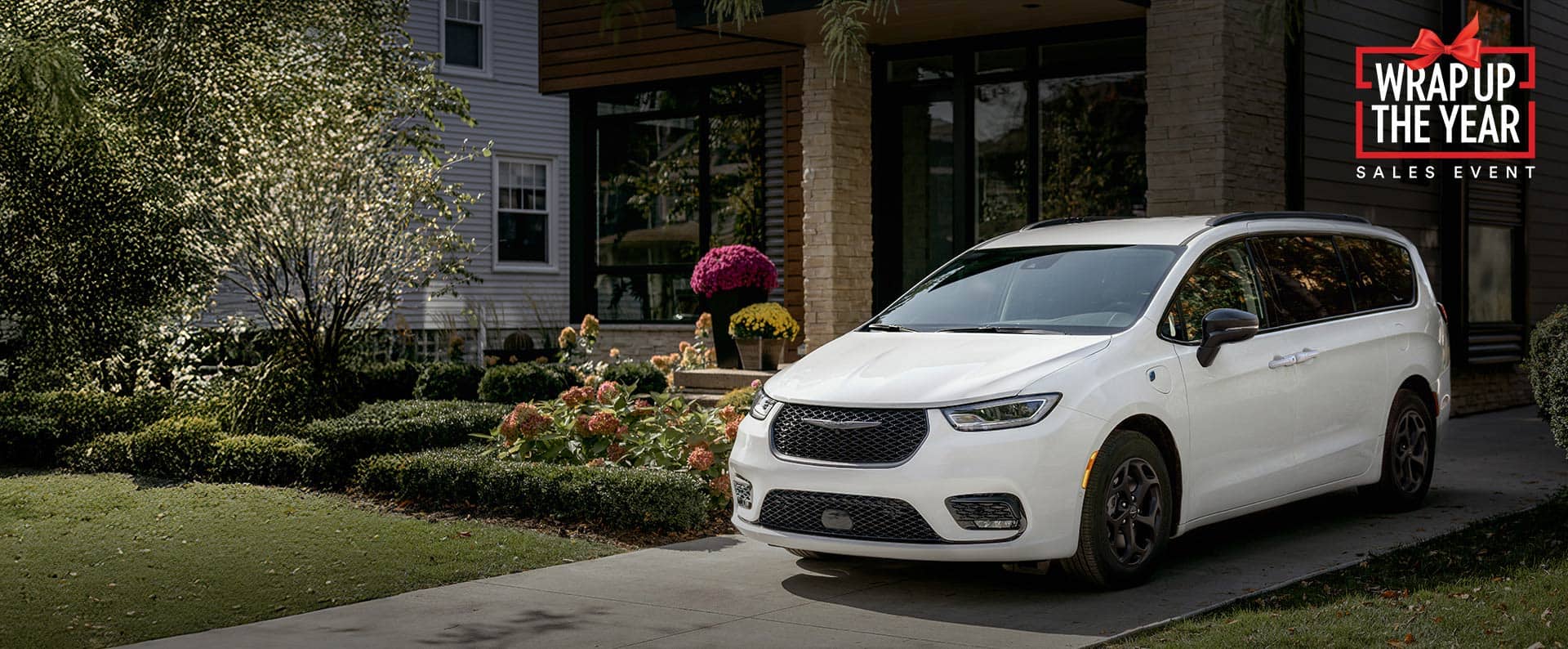 A driver-side front angle of a white 2025 Chrysler Pacifica Select Plug-in Hybrid with the Premium S Appearance Package, parked in the driveway of a neighborhood home. Wrap Up the Year Sales Event.