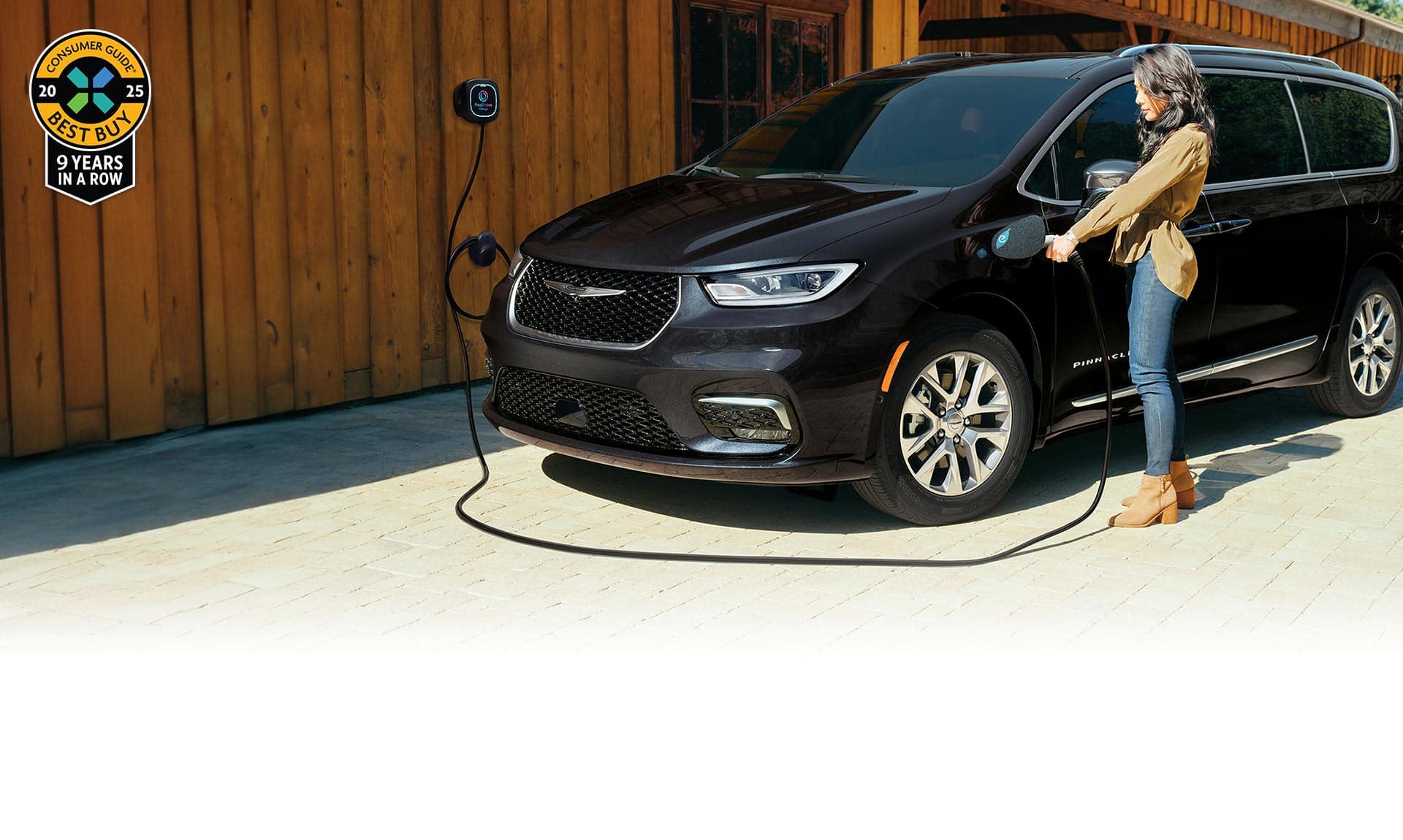 A driver-side front angle of a black 2025 Chrysler Pacifica Pinnacle Plug-in Hybrid parked outside a building with a charging station. A woman is plugging the station's charging cord into the vehicle's charging port. 2025 Consumer Guide Best Buy award.