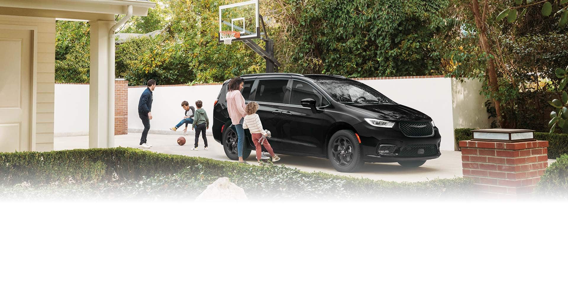 A black 2025 Chrysler Pacifica Select Plug-in Hybrid with the Premium S Appearance Package, parked in the driveway of a neighborhood home as the dad and two boys play basketball. The mom and daughter stand beside the hands-free power sliding side door as they each swing a foot under the door to open it.