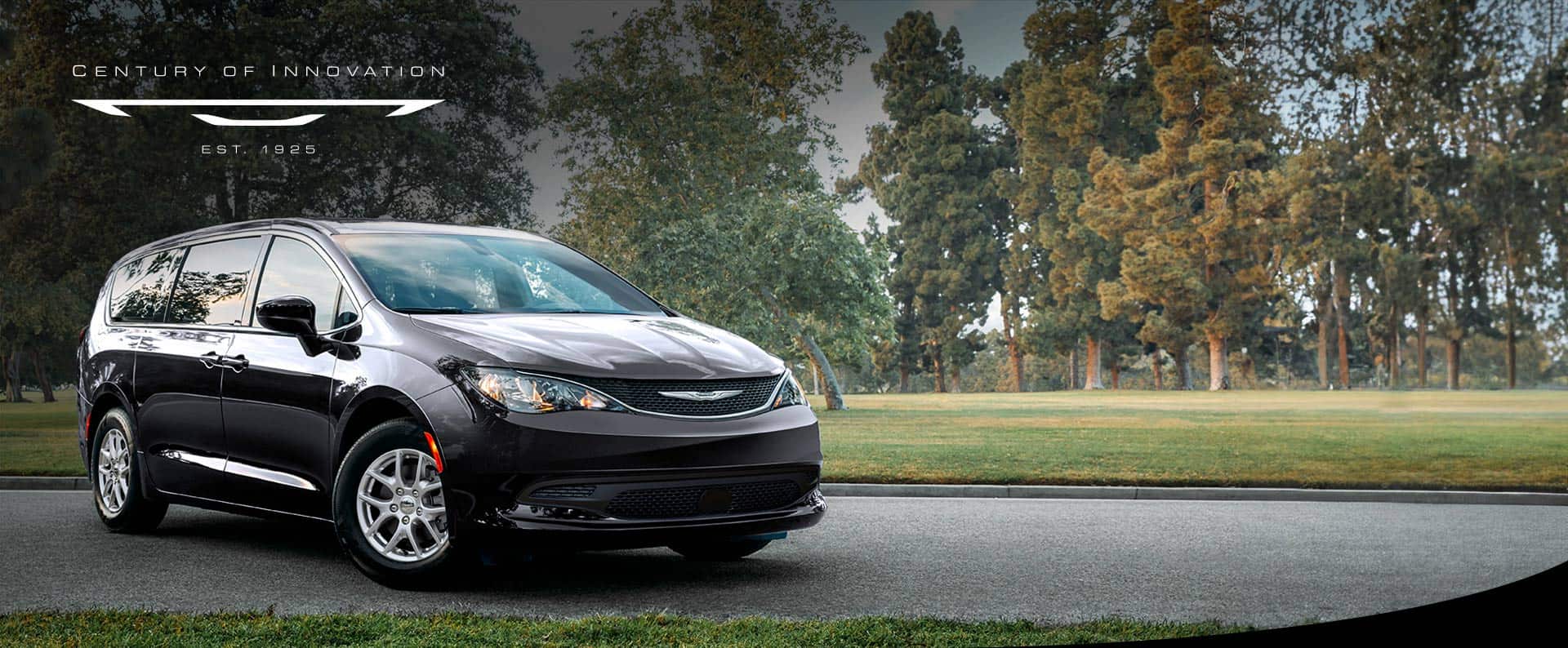A front pasengers side view of a black 2025 Chrysler Voyager LX in a parking lot of a park. Century of Innovation.