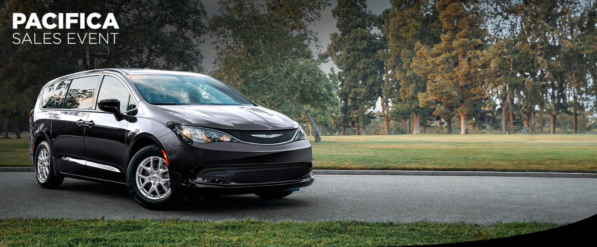 A front pasengers side view of a black 2025 Chrysler Voyager LX in a parking lot of a park. Chrysler Pacifica sales event.