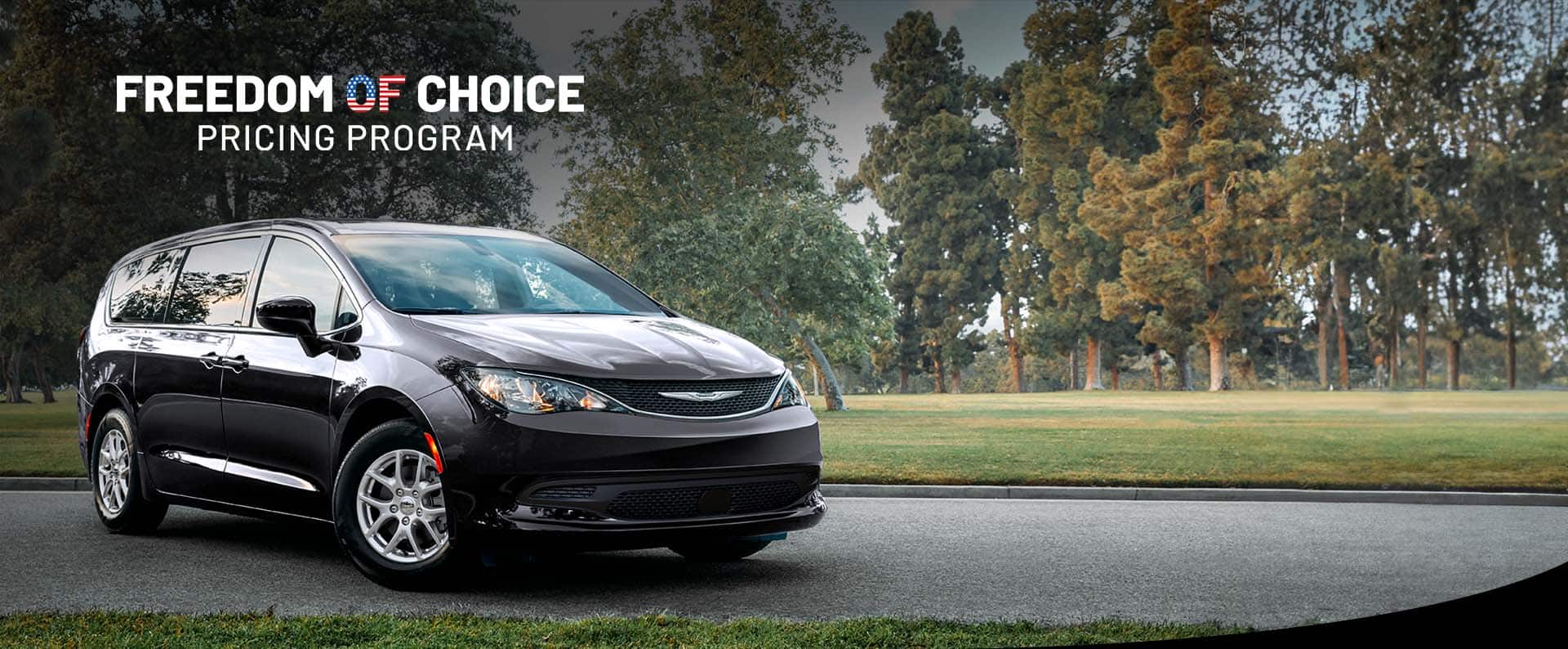 A front pasengers side view of a black 2025 Chrysler Voyager LX in a parking lot of a park. Freedom of Choice sales event.