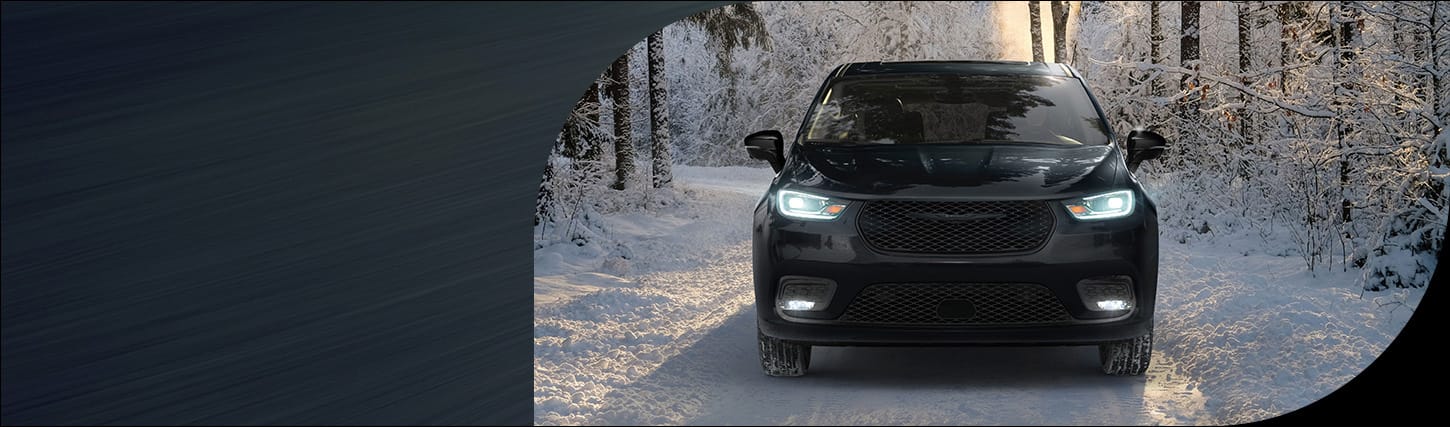 A head-on angle of a black 2026 Chrysler Pacifica Limited with its headlamps on, traveling down a snow-covered road in the woods.