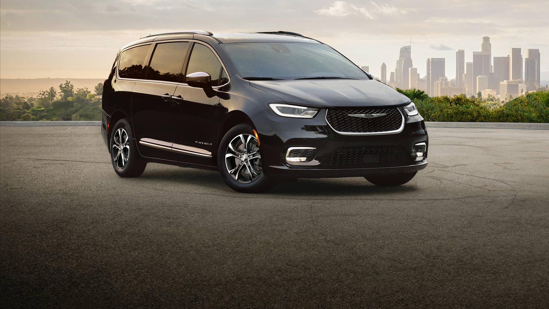 A passenger-side front angle of a black 2026 Chrysler Pacifica Pinnacle parked on a lot with a metropolitan skyline in the background.