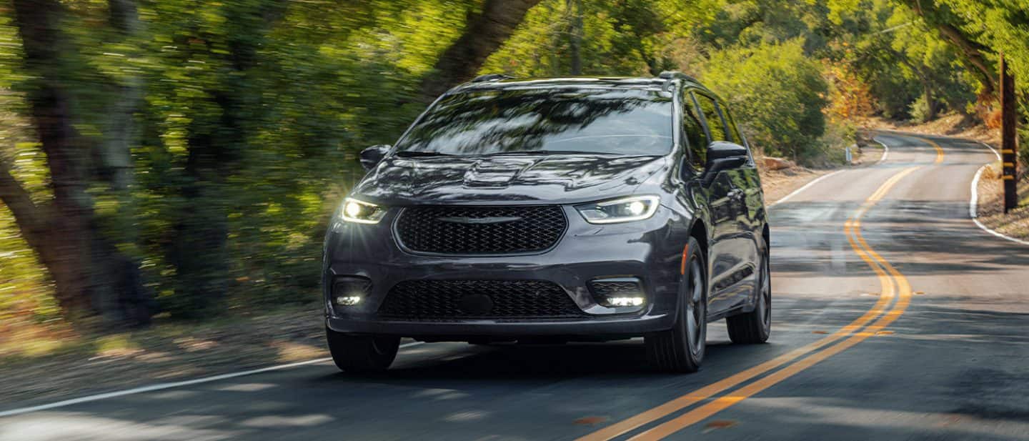 Display A gray 2026 Chrysler Pacifica Limited traveling down a winding highway in the woods.