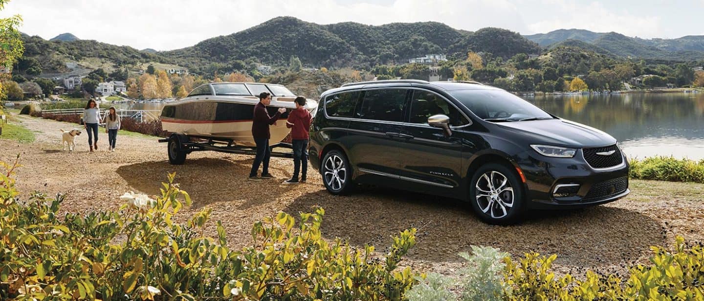 Display A black 2026 Chrysler Pacifica Pinnacle  parked beside a lake, towing a motorboat, with a family of four nearby.