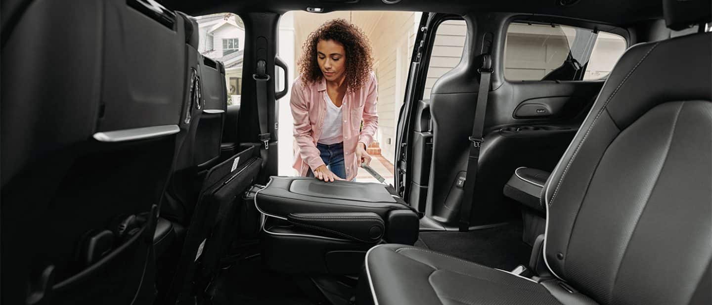 Display The interior of the 2026 Chrysler Pacifica with a woman at the open passenger-side sliding door. She is folding one of the second-row seats into the floor.