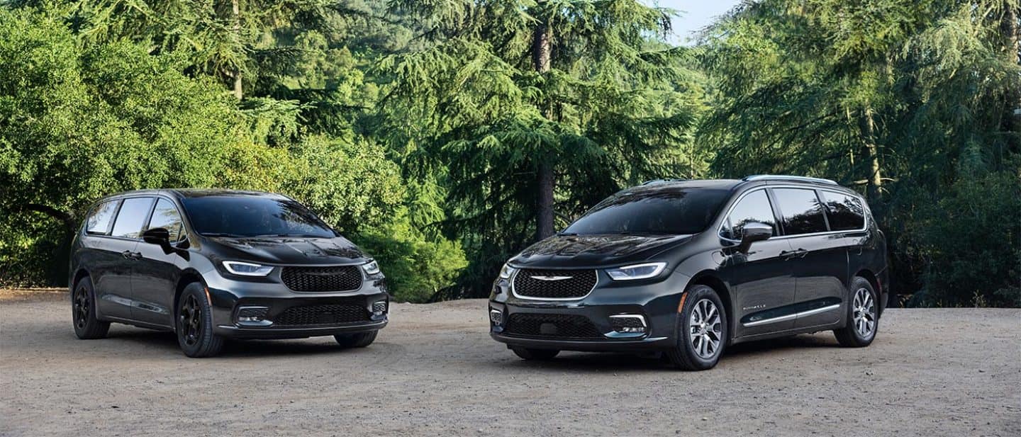 Display Two blue 2026 Chrysler Pacifica Plug-in Hybrid models parked perpendicular to each other, at a park.  On the left, a Pacifica Plug-in Hybrid Select with the Premium S Appearance Package, and on the right, a Pacifica Plug-in Hybrid Pinnacle.