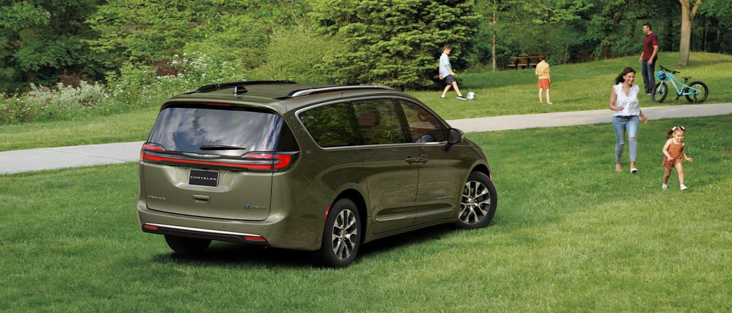 Display A passenger-side rear angle of a 2026 Chrysler Pacifica Plug-in Hybrid parked on the grass at a park with children and adults nearby.