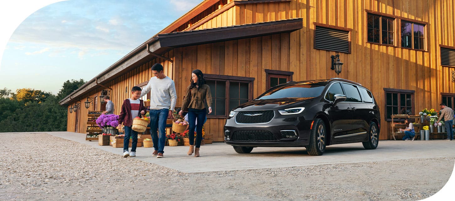 A black 2026 Chrysler Pacifica Plug-In Hybrid Pinnacle parked at a farmers market, as a family of three walk nearby.