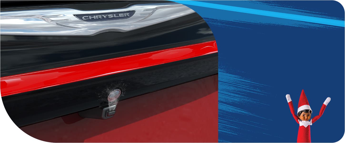 A close-up of the rear camera washer spraying cleaner onto the camera lens of a 2026 Chrysler Pacifica, as Sparkles, the Elf on the Shelf happily stands in the lower right corner of the image.