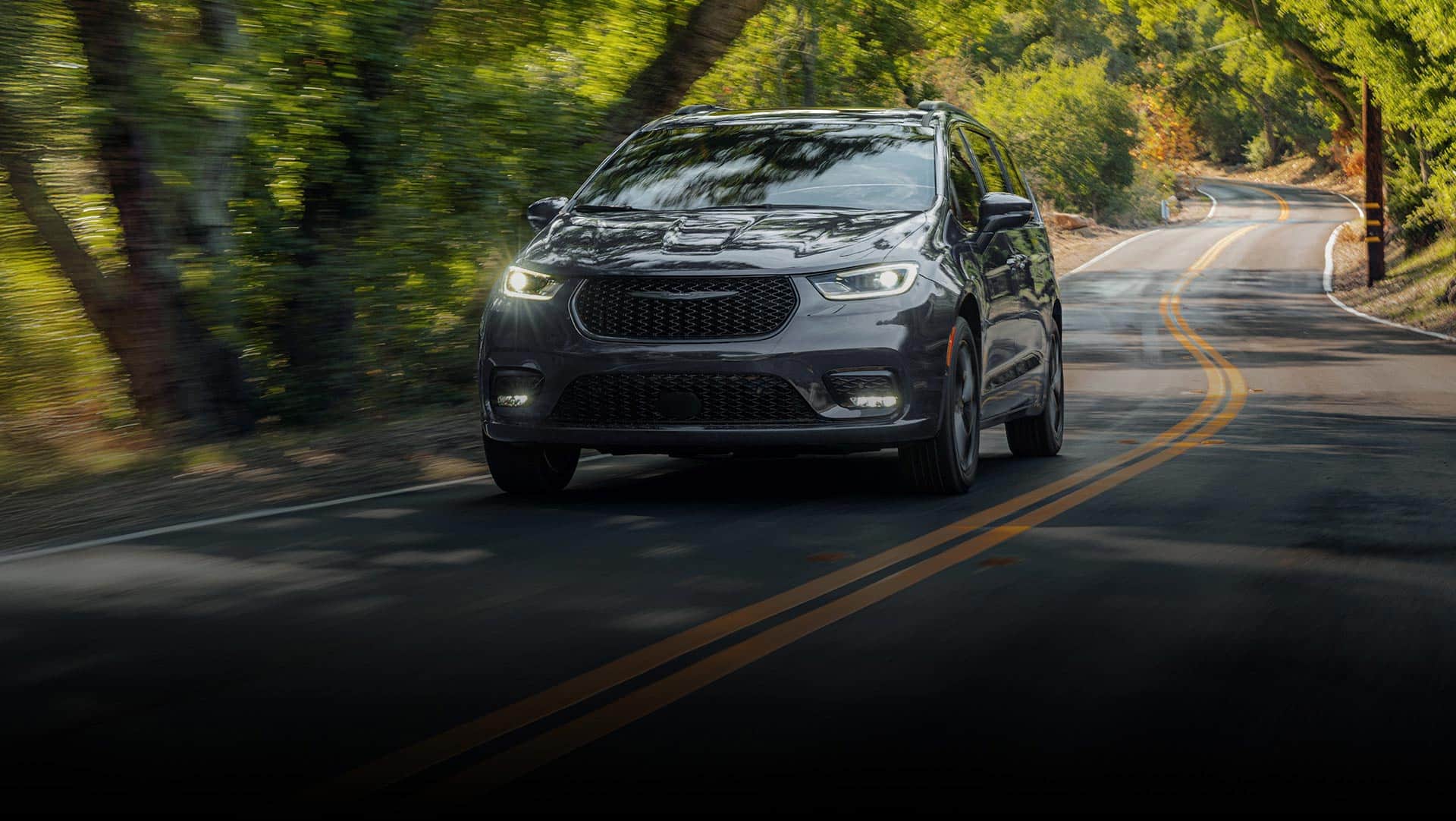 A gray 2026 Chrysler Pacifica Limited traveling down a winding highway in the woods.