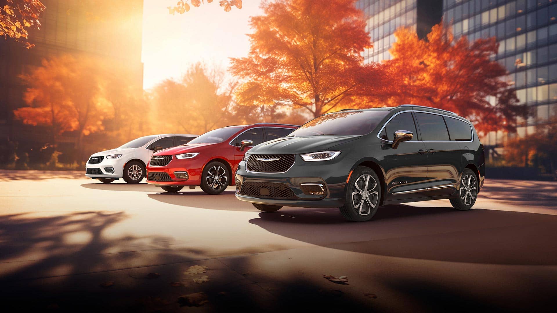 Three 2026 Chrysler minivan models parked on an oceanside beach. From left to right: a white Chrysler Voyager, a blue Pacifica Pinnacle Plug-in Hybrid and a red Pacifica Pinnacle.