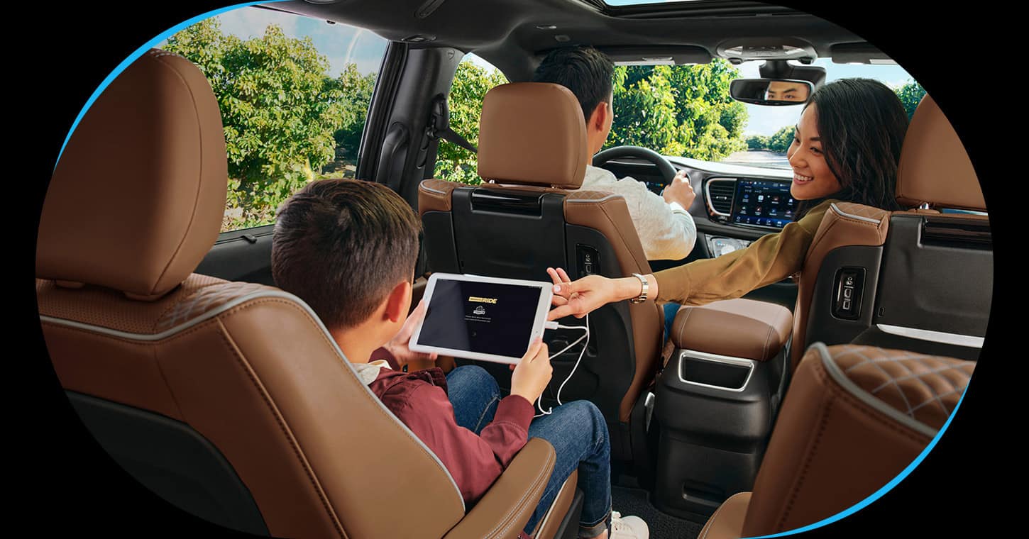 The interior of the 2026 Chrysler Pacifica Pinnacle with a man driving, a woman reaching into the rear and a boy in the second row holding a tablet that is plugged into an outlet.