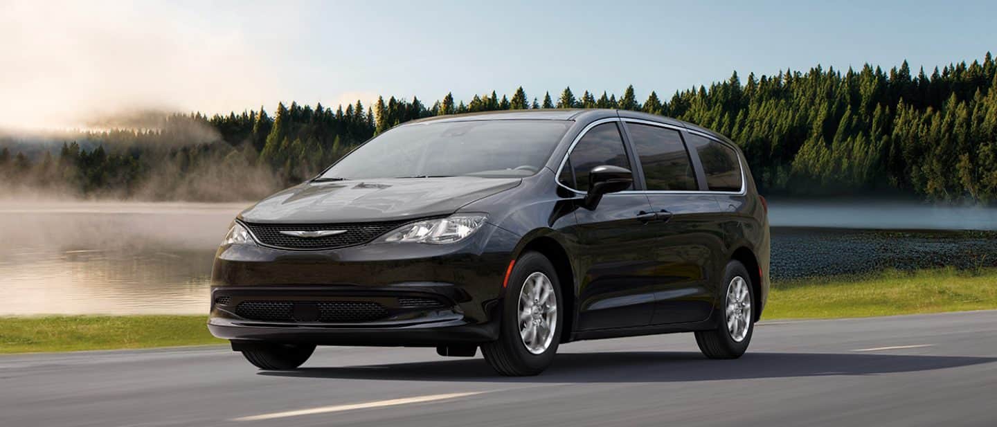 A driver-side front angle of a black 2026 Chrysler Voyager LX traveling on a lakeside highway.