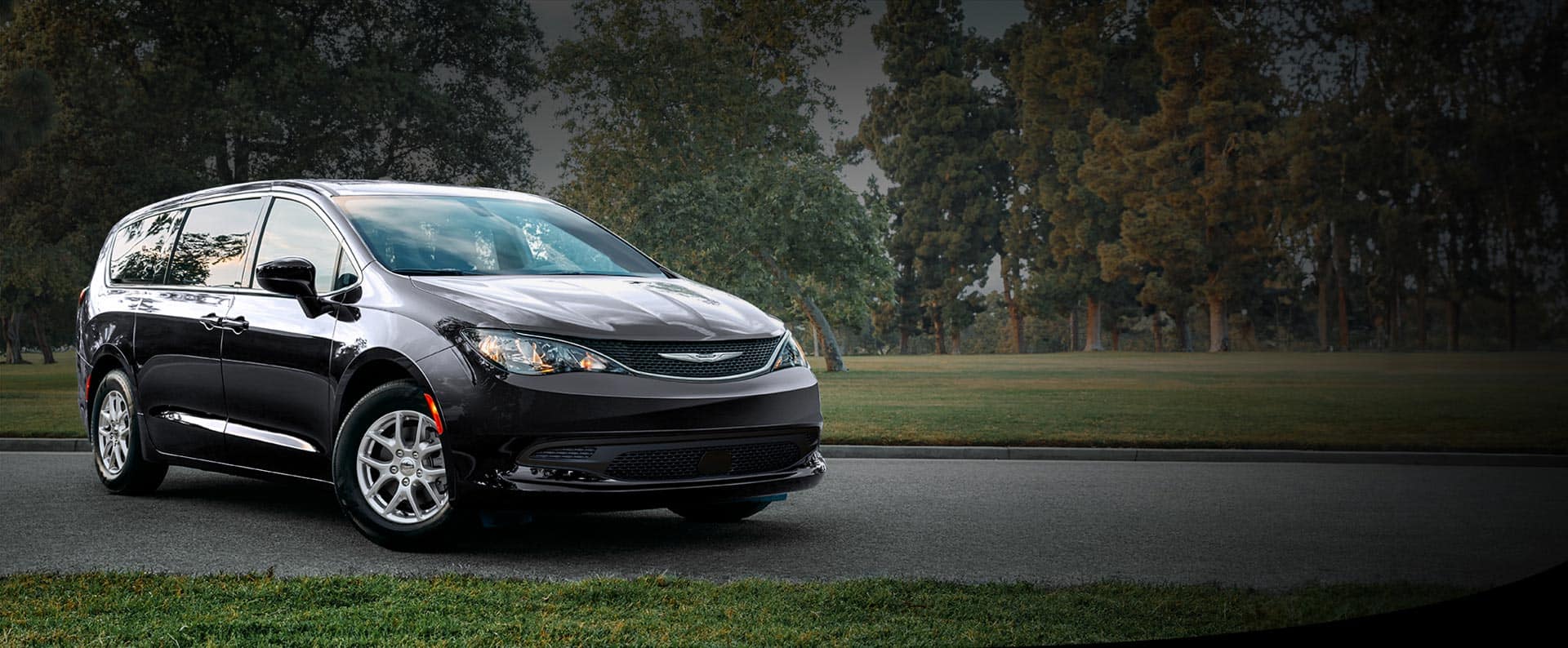 A front passengers side view of a black 2026 Chrysler Voyager LX in a parking lot of a park.
