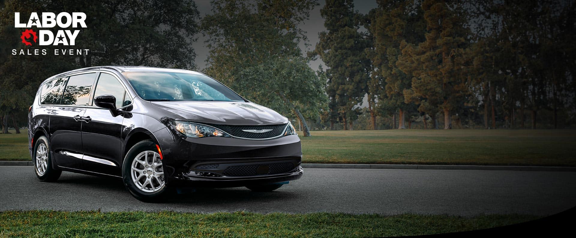 A front passengers side view of a black 2026 Chrysler Voyager LX in a parking lot of a park. Labor day Sales Event.