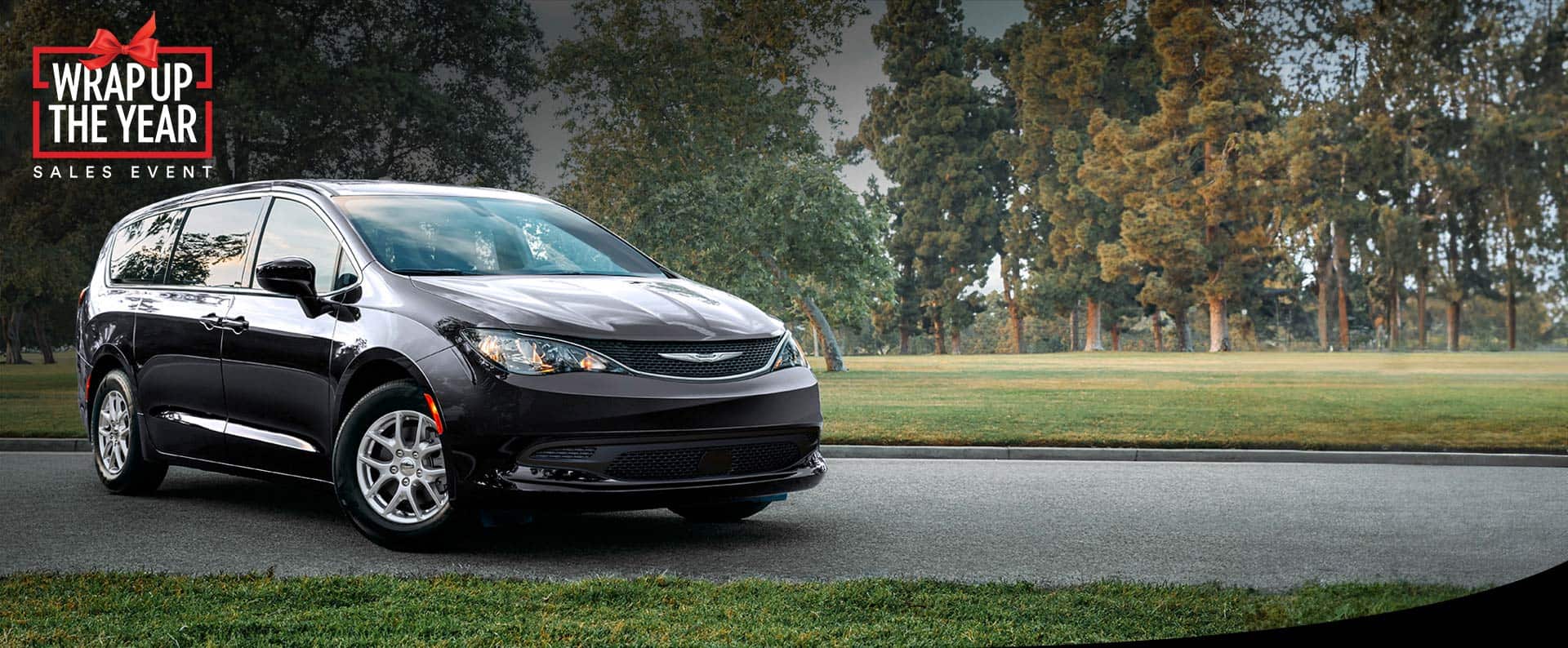 A front passengers side view of a black 2025 Chrysler Voyager LX in a parking lot of a park. Wrap Up the Year Sales Event.