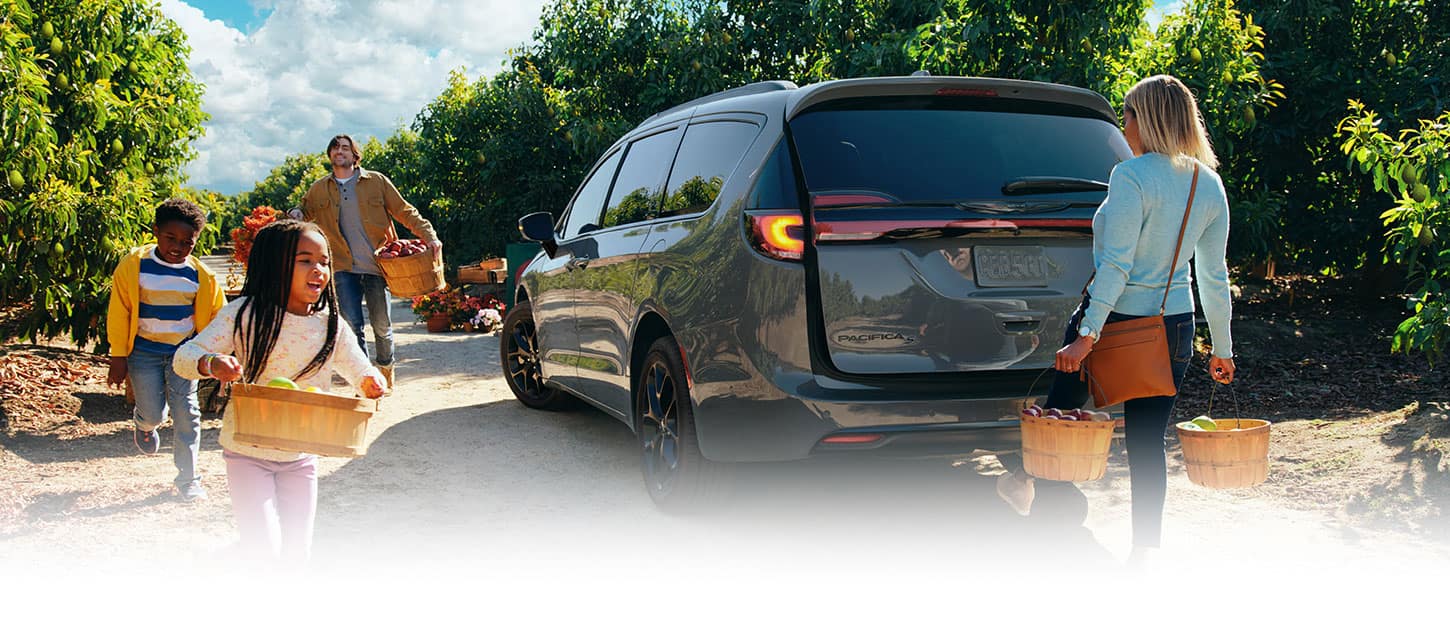 The gray 2023 Chrysler Pacifica Limited with S Appearance Package parked in an orchard. A mother carrying baskets of fruit in both hands swings her foot under the rear bumper as the power liftgate begins to open. The father and two children carry bushels of produce toward the vehicle.