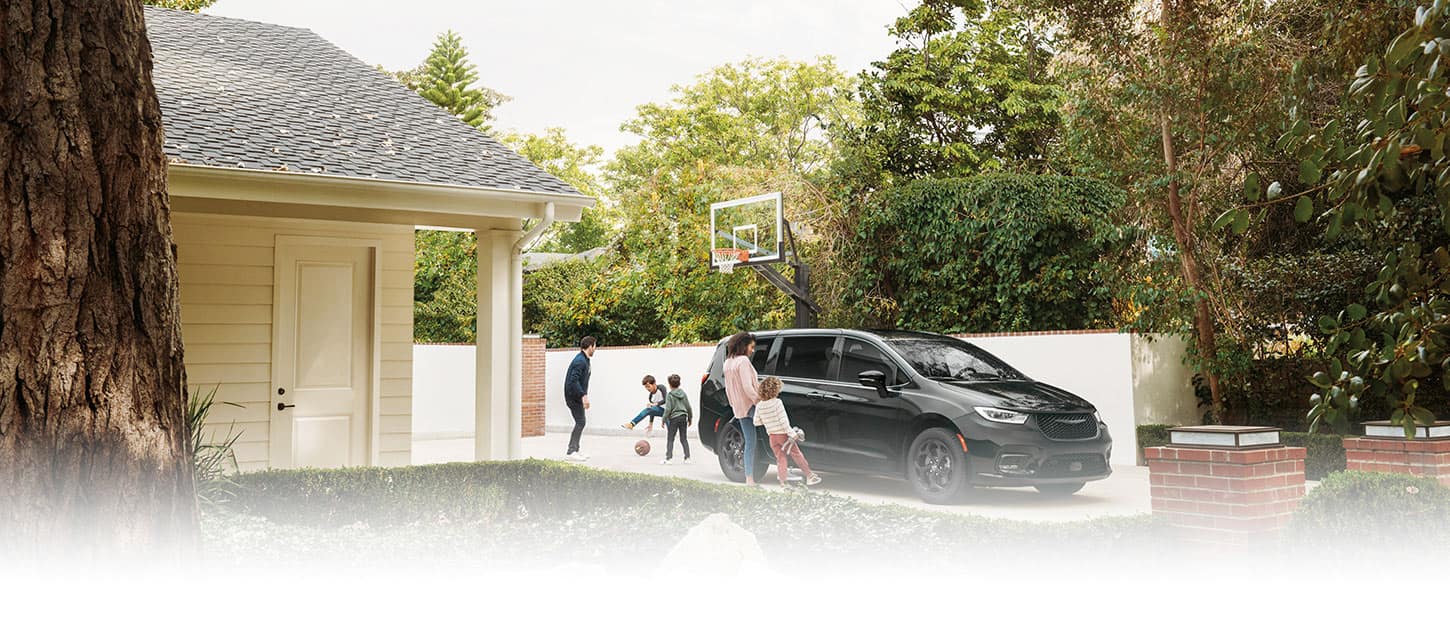 A gray 2023 Chrysler Pacifica Limited with S Appearance Package Plug-In Hybrid parked in the driveway of a suburban home with a mother and daughter opening the handsfree power sliding side door while the father and two sons play basketball in the driveway.