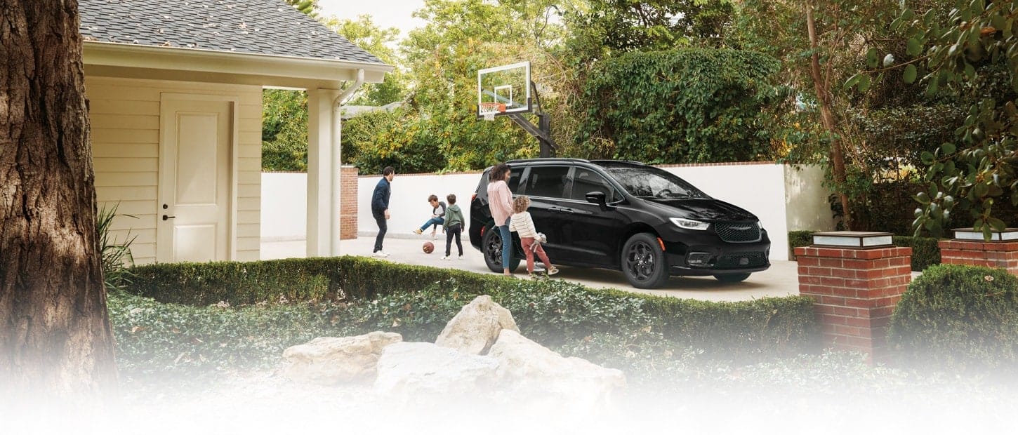 A gray 2023 Chrysler Pacifica Limited with S Appearance Package Plug-In Hybrid parked in the driveway of a suburban home with a mother and daughter opening the handsfree power sliding side door while the father and two sons play basketball in the driveway.