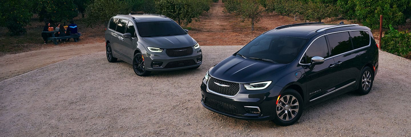 Two 2021 Chrysler Pacifica models: the Limited with the S Appearance Package and the Pinnacle Hybrid, parked in a gravel clearing amidst trees planted in rows.