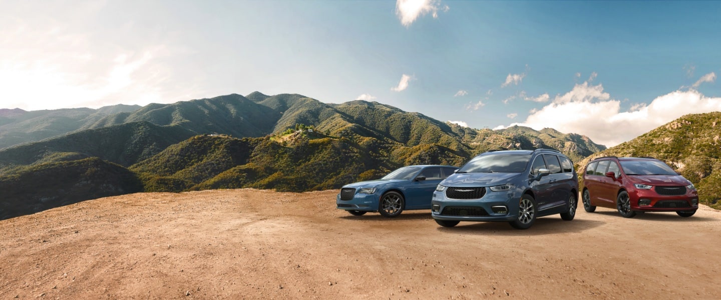 The 2022 Chrysler Brand lineup, parked on a clearing in the mountains. From left to right: a blue 2022 Chrysler 300 Touring L, a blue 2022 Chrysler Pacifica Pinnacle Hybrid and a 2022 red Pacifica Limited with S Appearance Package.