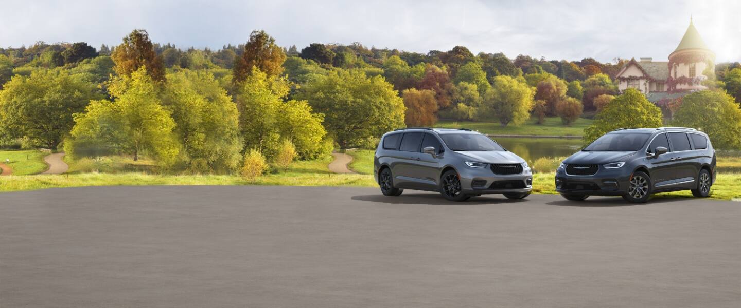 A 2023 Chrysler Pacifica Limited with S Appearance Package (left) and 2023 Chrysler Pacifica Pinnacle (right) parked beside a pond with a castle-like home in the distance.