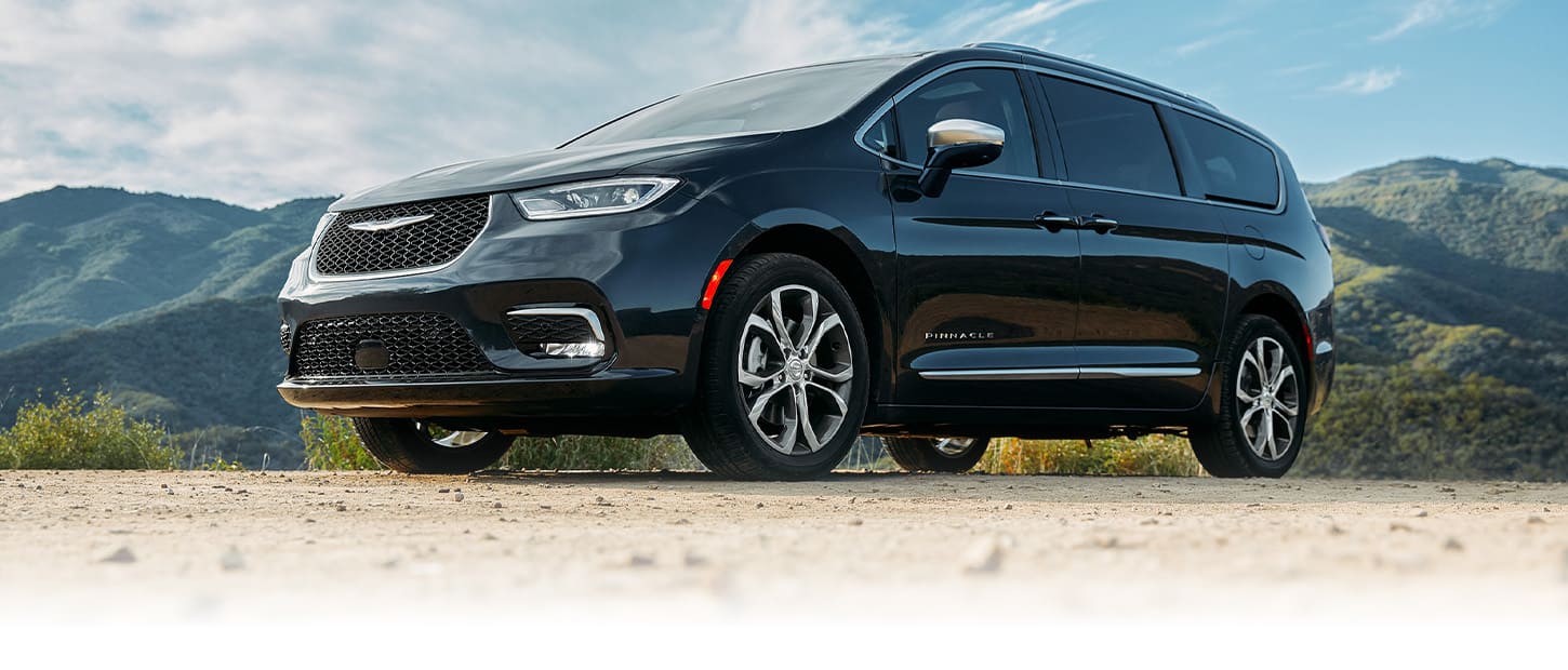 An angled driver-side profile of a black 2024 Chrysler Pacifica Pinnacle parked on a clearing with mountains in the background.