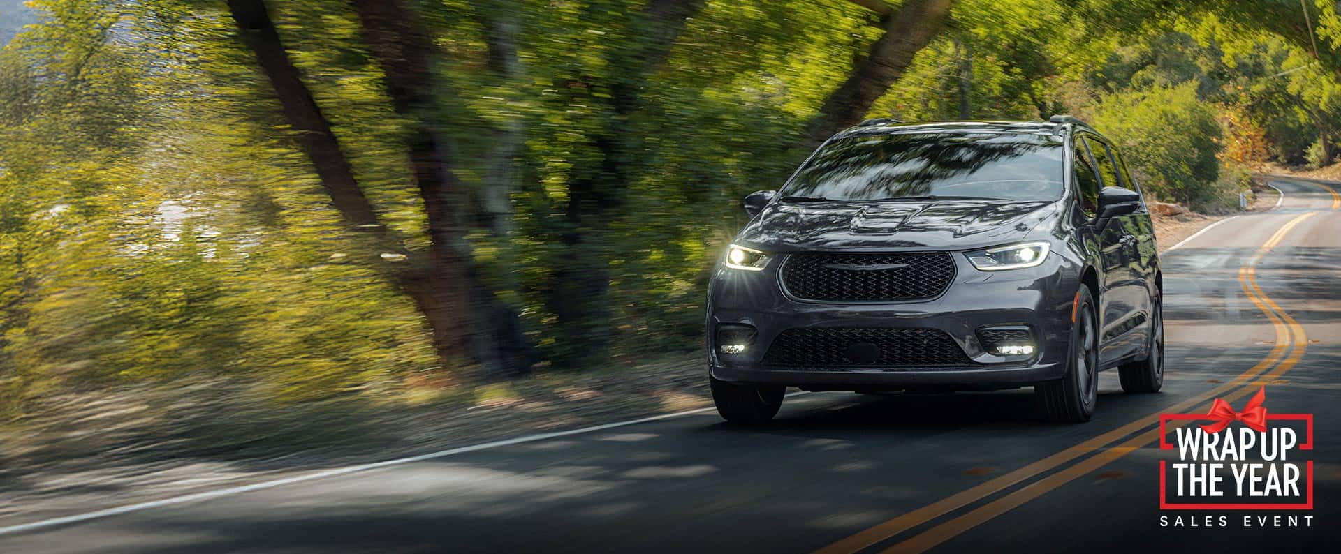 A gray 2026 Chrysler Pacifica Limited traveling down a winding highway in the woods. Wrap Up the Year Sales Event.