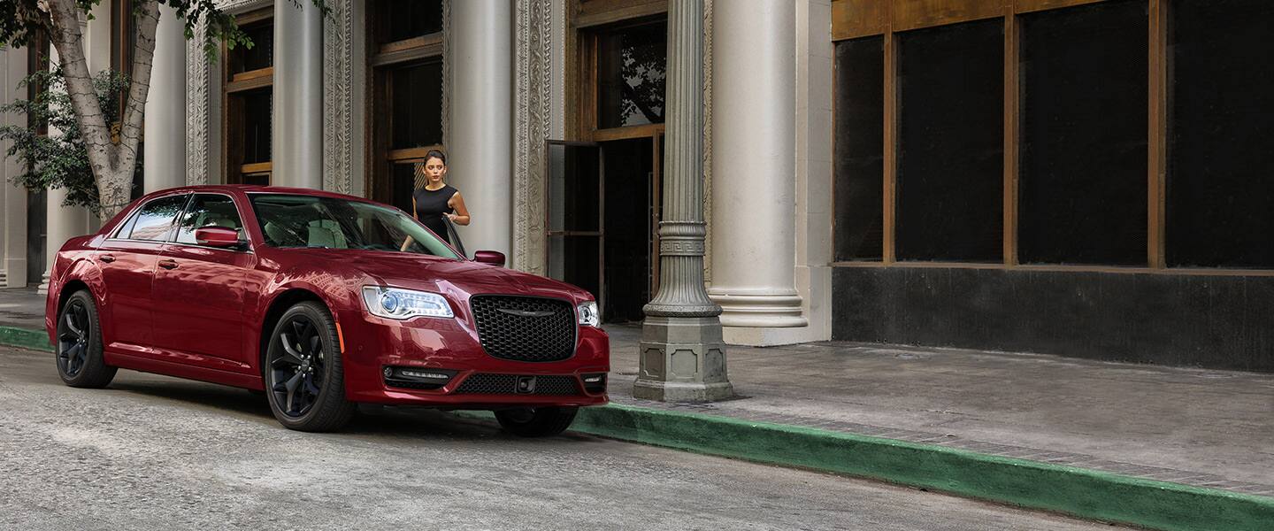 A red 2023 Chrysler 300 Touring L parked beside a commercial building with a woman standing beside the open driver-side door. 