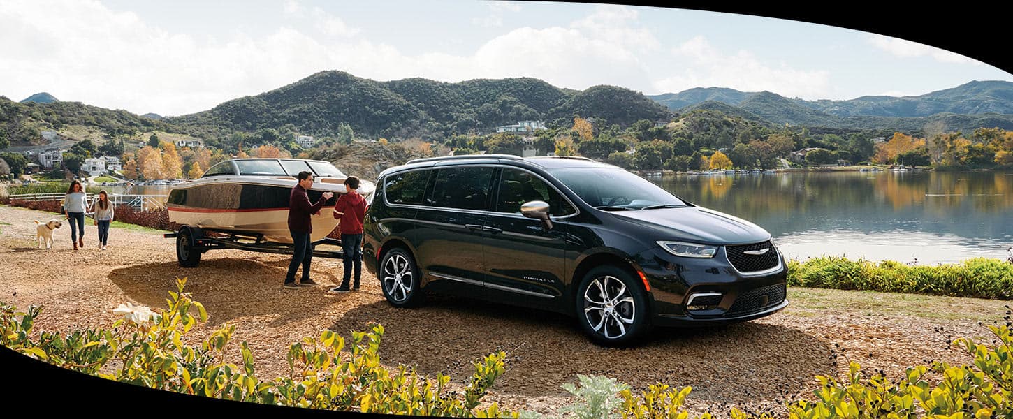 A black 2024 Chrysler Pacifica Pinnacle parked beside a lake, towing a motorboat, with a family of four nearby.