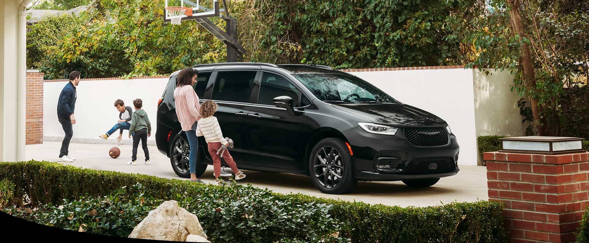 A black 2025 Chrysler Pacifica Limited parked in the driveway of a neighborhood home. A mom and daughter stand beside the hands-free power sliding side door as they each swing a foot under the door to open it.