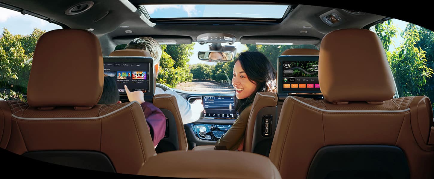 The front passenger in the 2025 Chrysler Pacifica Pinnacle turning to look at a child in the second row who is browsing options on the front seatback-mounted touchscreen.