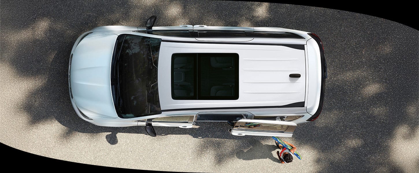 A slider that when pulled to the right reveals an overhead angle of a white 2025 Chrysler Pacifica with a sunroof and a child walking toward the driver-side sliding door. When pulled to the left the slider reveals the same Chrysler Pacifica, but with its entire roof removed, revealing the interior full with a driver and front passenger, a child in the second row, two children in the third row and cargo behind the third-row seat.