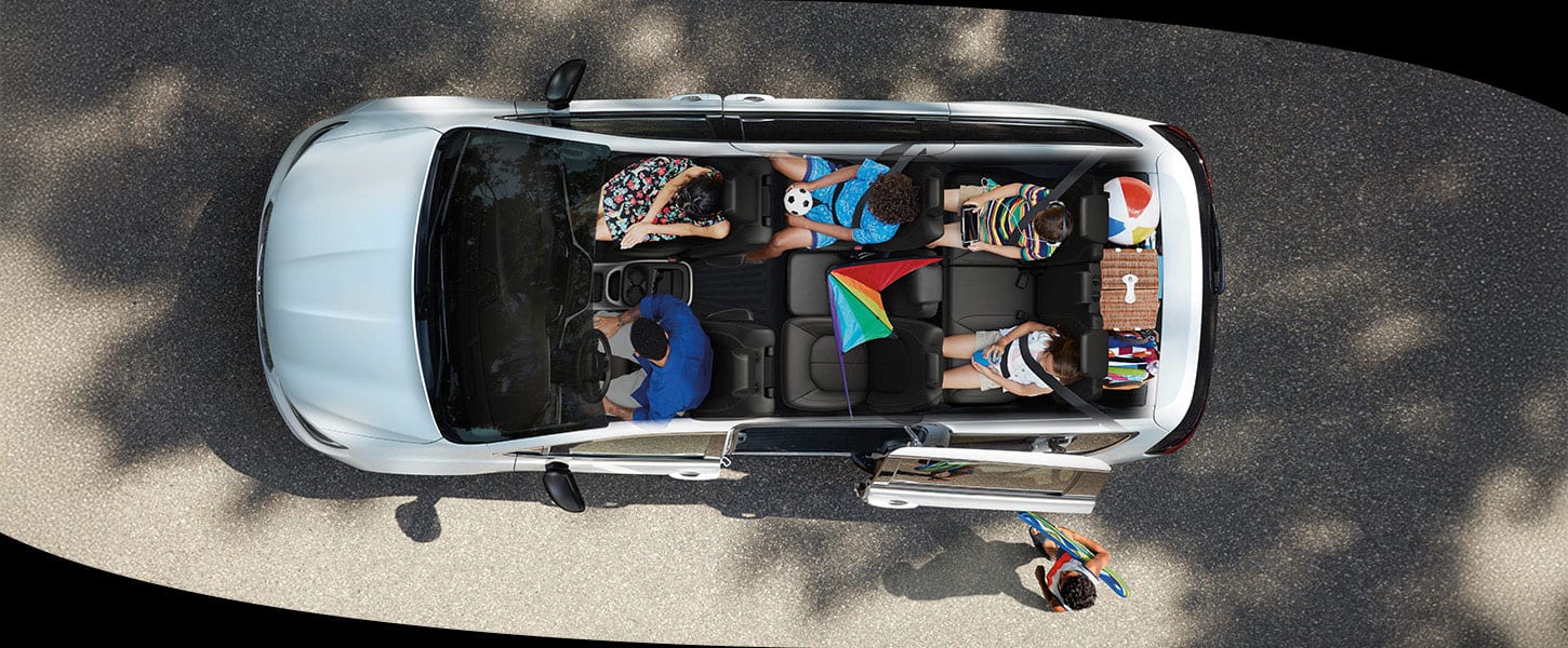 A slider that when pulled to the left reveals an overhead angle of a white 2025 Chrysler Pacifica with its entire roof removed, revealing the interior full with a driver and front passenger, a child in the second row, two children in the third row, cargo behind the third-row seat and a child outside the vehicle, walking toward the driver-side sliding door. When pulled to the right the slider reveals the same overhead angle of the Chrysler Pacifica exterior with sunroof.
