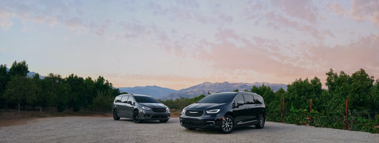 Two 2021 Chrysler Pacifica models: the Limited with the S Appearance Package and the Pinnacle Hybrid, parked in a gravel clearing amidst trees planted in rows.