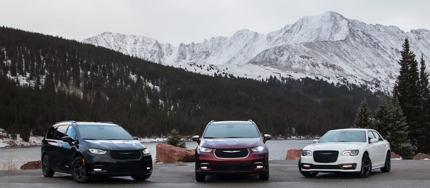 The 2022 Chrysler Brand lineup, from left to right: the Chrysler Pacifica Hybrid Limited with the S Appearance Package, the Chrysler Pacifica Pinnacle and the Chrysler 300S, all parked beside a lake with majestic mountains in the background.